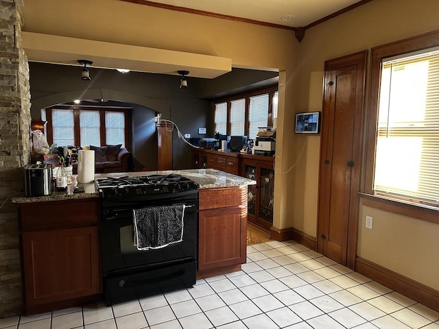 kitchen with light tile patterned floors, ornamental molding, brown cabinets, dark stone countertops, and gas stove