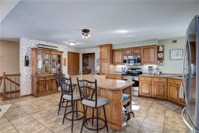 kitchen with wallpapered walls, visible vents, appliances with stainless steel finishes, a kitchen breakfast bar, and open shelves