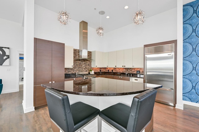 kitchen with a towering ceiling, stainless steel built in fridge, wall chimney exhaust hood, light wood finished floors, and an inviting chandelier