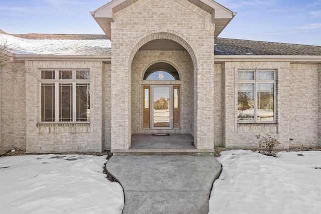 snow covered property entrance with brick siding and roof with shingles