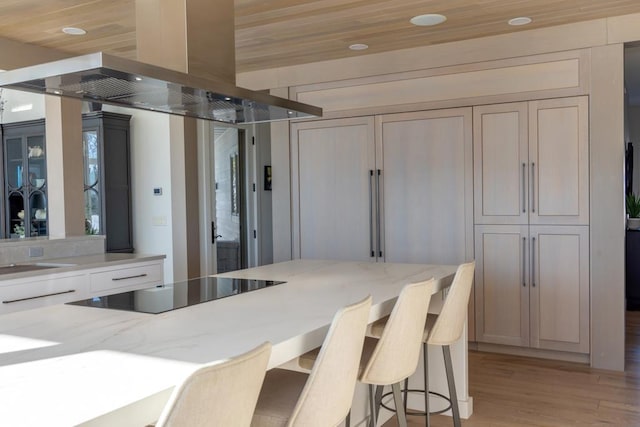 kitchen featuring black electric stovetop, light stone counters, a kitchen breakfast bar, light wood-style floors, and island exhaust hood