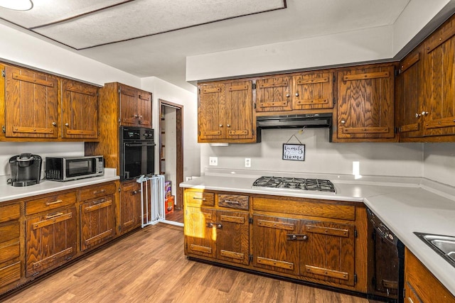 kitchen featuring under cabinet range hood, light countertops, black appliances, light wood finished floors, and brown cabinetry