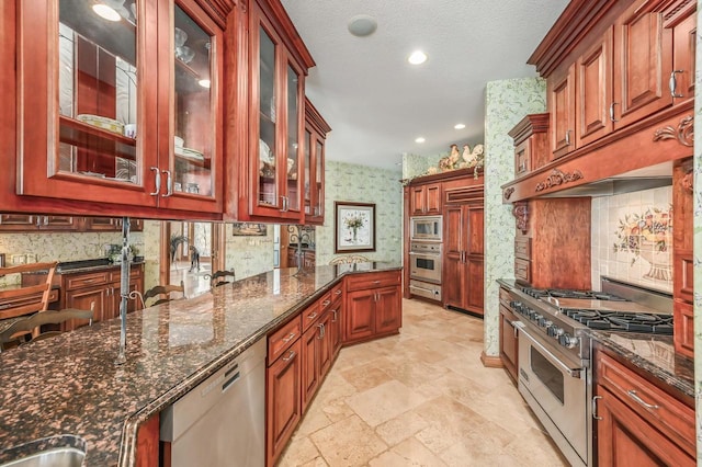 kitchen featuring stone tile floors, wallpapered walls, dark stone counters, stainless steel appliances, and glass insert cabinets