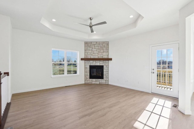 unfurnished living room featuring light wood finished floors, a stone fireplace, a tray ceiling, and a healthy amount of sunlight