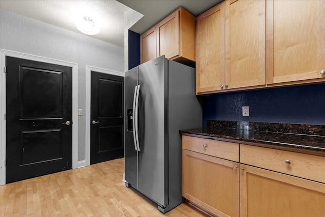 kitchen with dark stone counters, a textured wall, stainless steel fridge with ice dispenser, light brown cabinetry, and light wood-type flooring