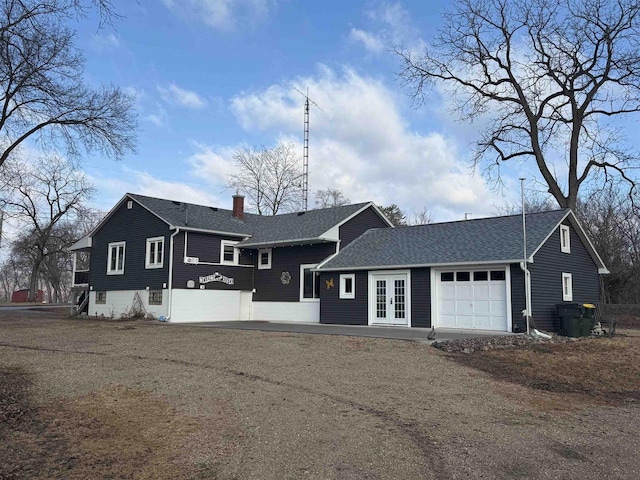 view of front facade with a garage, dirt driveway, a chimney, roof with shingles, and french doors