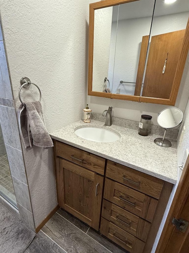 bathroom with a textured wall, wood finish floors, tiled shower, and vanity