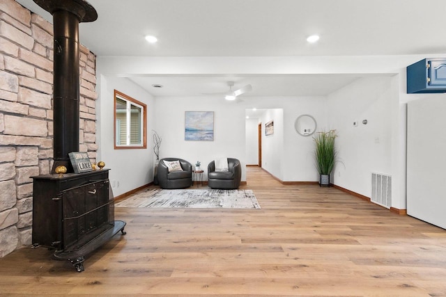 sitting room with visible vents, a ceiling fan, a wood stove, light wood-type flooring, and baseboards