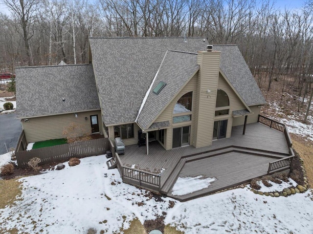 snow covered back of property with a deck, roof with shingles, and a chimney