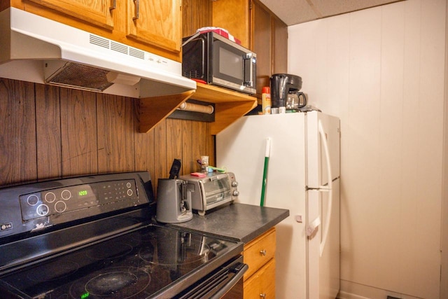 kitchen featuring brown cabinets, dark countertops, appliances with stainless steel finishes, wooden walls, and under cabinet range hood