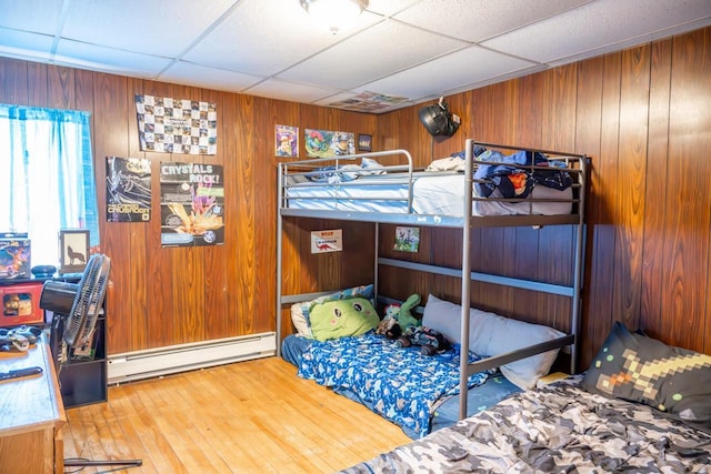 bedroom with a drop ceiling, wood walls, baseboard heating, and hardwood / wood-style flooring