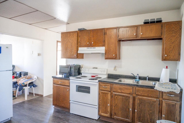 kitchen with white appliances, brown cabinetry, dark countertops, under cabinet range hood, and a sink