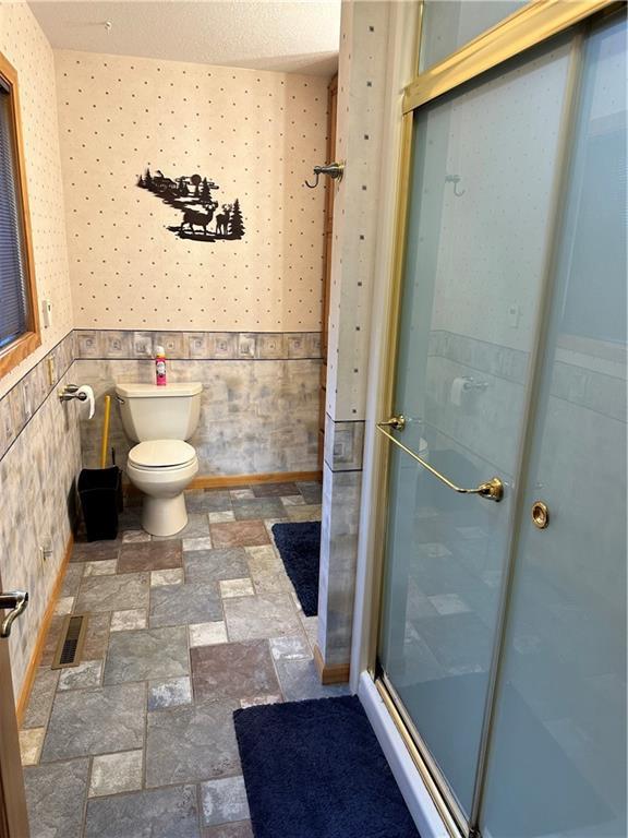 bathroom featuring toilet, wallpapered walls, a shower stall, and a wainscoted wall