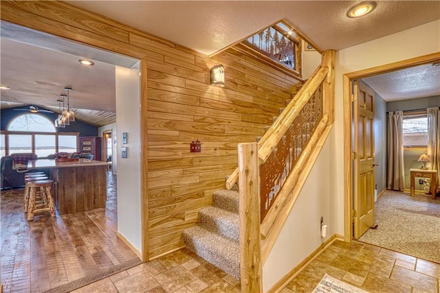 stairway featuring a textured ceiling, wood walls, baseboards, vaulted ceiling, and stone tile flooring