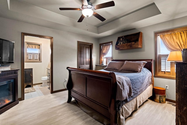 bedroom with a tray ceiling, a glass covered fireplace, light wood-style flooring, and baseboards