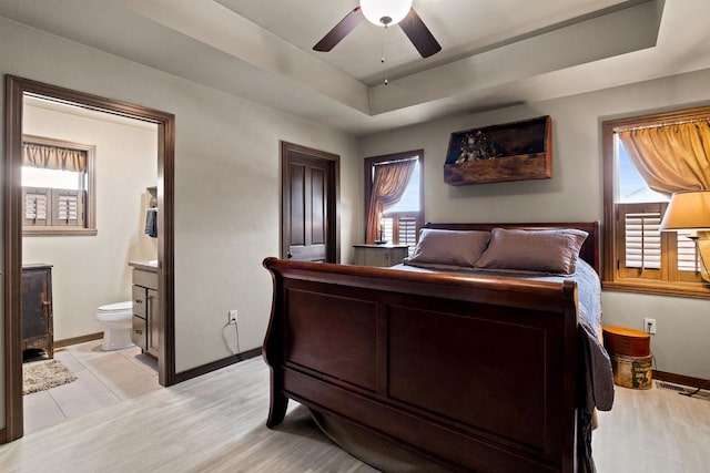 bedroom featuring connected bathroom, a ceiling fan, baseboards, light wood-type flooring, and a tray ceiling