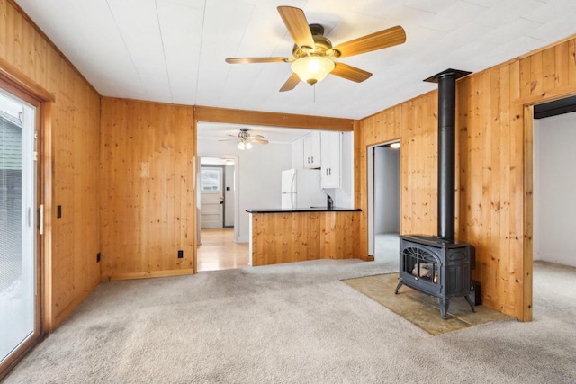 unfurnished living room with a ceiling fan, a wood stove, light carpet, and wooden walls