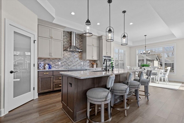 kitchen featuring dark wood-style flooring, wall chimney exhaust hood, a tray ceiling, an island with sink, and tasteful backsplash