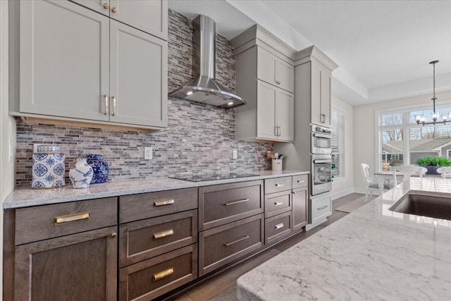 kitchen featuring wall chimney exhaust hood, decorative backsplash, light stone countertops, and an inviting chandelier