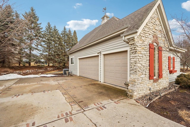 view of side of property with central air condition unit, a garage, stone siding, driveway, and a chimney