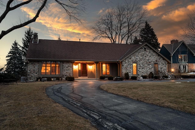 view of front of house with stone siding, a chimney, curved driveway, and a yard