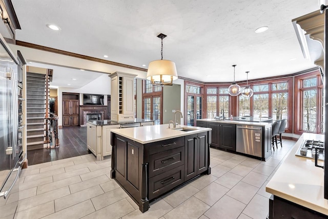 kitchen with a kitchen island with sink, stainless steel appliances, a fireplace, a sink, and decorative columns