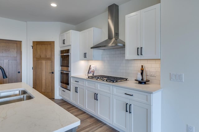 kitchen with light wood finished floors, appliances with stainless steel finishes, wall chimney range hood, white cabinetry, and a sink