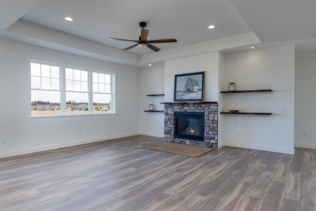 unfurnished living room with baseboards, a raised ceiling, wood finished floors, a stone fireplace, and recessed lighting