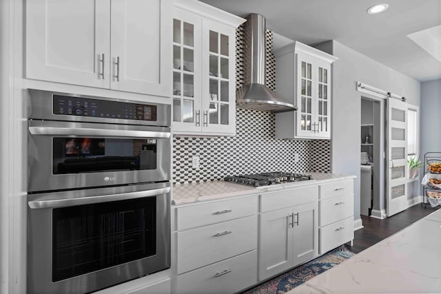 kitchen featuring light stone counters, a barn door, appliances with stainless steel finishes, wall chimney range hood, and decorative backsplash