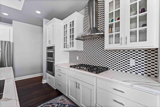 kitchen with dark wood-type flooring, appliances with stainless steel finishes, white cabinets, wall chimney range hood, and glass insert cabinets