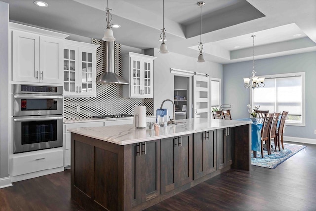 kitchen featuring a tray ceiling, a barn door, gas stovetop, double oven, and wall chimney range hood