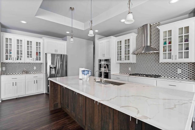 kitchen with a sink, dark wood-style floors, stainless steel appliances, wall chimney exhaust hood, and a raised ceiling