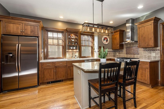 kitchen featuring light wood finished floors, decorative backsplash, appliances with stainless steel finishes, wall chimney exhaust hood, and brown cabinets