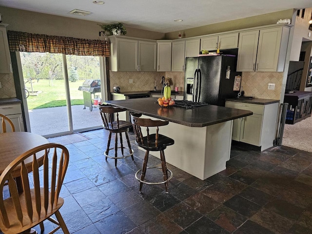 kitchen featuring stone tile floors, decorative backsplash, black fridge with ice dispenser, dark countertops, and a kitchen breakfast bar