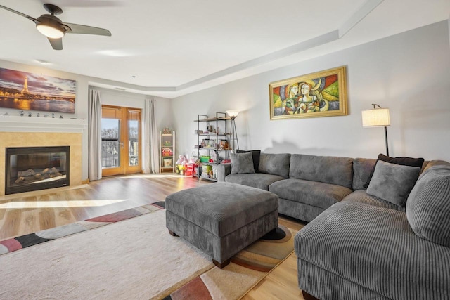 living room featuring a raised ceiling, a ceiling fan, a glass covered fireplace, wood finished floors, and french doors