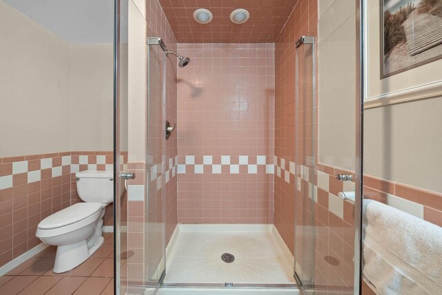 bathroom with a wainscoted wall, tile walls, toilet, a shower stall, and tile patterned flooring