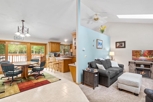 living area featuring vaulted ceiling with skylight, a warm lit fireplace, light tile patterned flooring, and ceiling fan with notable chandelier