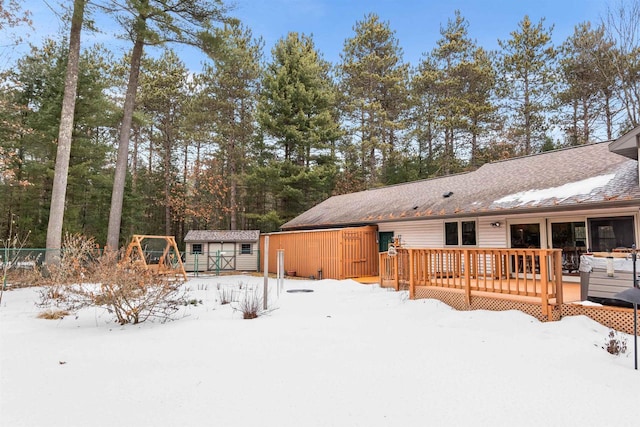 snow covered house with an outbuilding, a shingled roof, a storage shed, fence, and a wooden deck