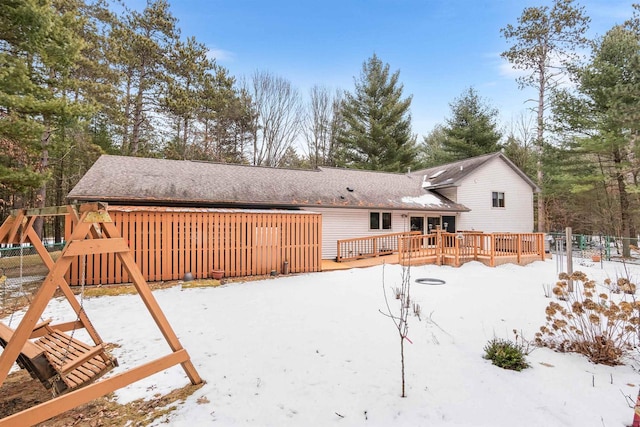 snow covered back of property with a shingled roof, fence, a deck, and a playground