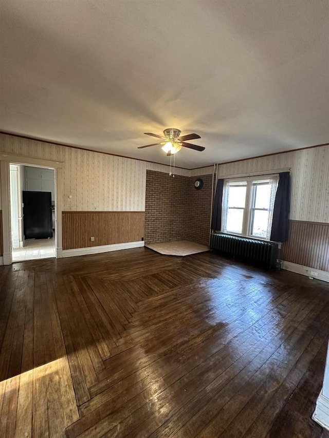 unfurnished living room with wallpapered walls, a ceiling fan, wainscoting, radiator, and wood-type flooring