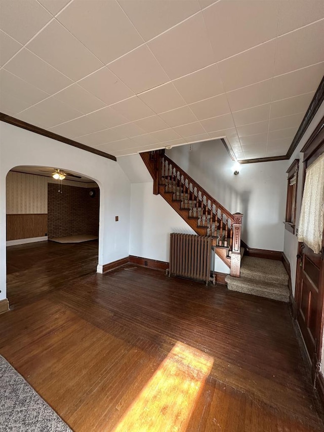 unfurnished living room with arched walkways, stairway, radiator heating unit, ornamental molding, and hardwood / wood-style floors