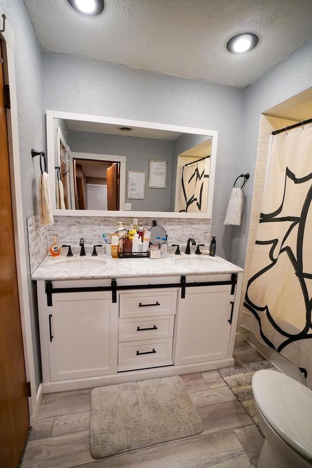 full bath with double vanity, backsplash, toilet, wood tiled floor, and a textured ceiling