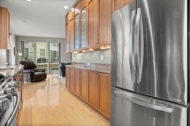 kitchen featuring light stone countertops, brown cabinetry, glass insert cabinets, appliances with stainless steel finishes, and light wood-type flooring