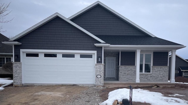 view of front of house with driveway, roof with shingles, a porch, an attached garage, and stone siding