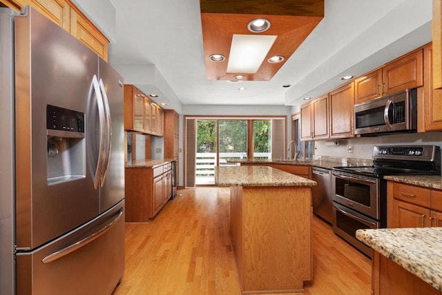 kitchen with light stone countertops, a center island, appliances with stainless steel finishes, light wood-type flooring, and brown cabinets