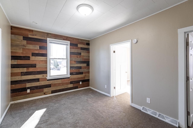 carpeted empty room featuring wooden walls, an accent wall, visible vents, baseboards, and ornamental molding