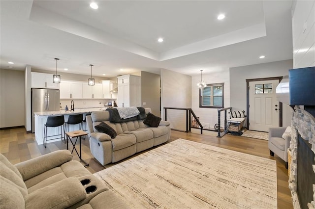 living room with a tray ceiling, a stone fireplace, light wood-style floors, a notable chandelier, and recessed lighting