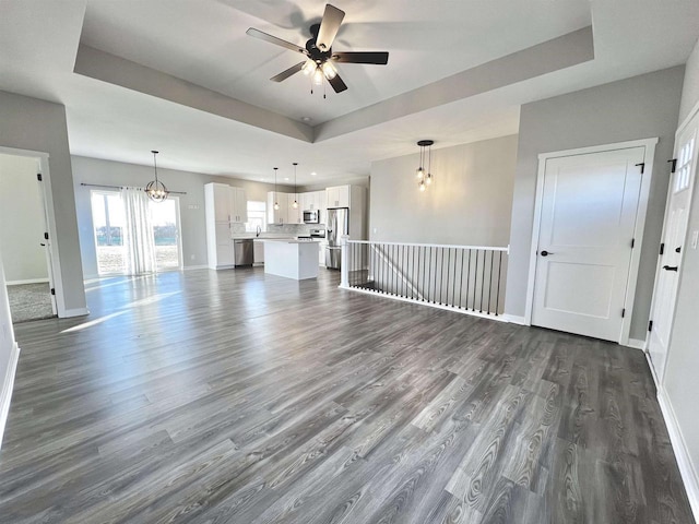 unfurnished living room featuring dark wood-style floors, a raised ceiling, baseboards, and ceiling fan with notable chandelier