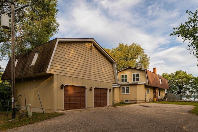 view of side of property featuring driveway, a shingled roof, a gambrel roof, a chimney, and an attached garage