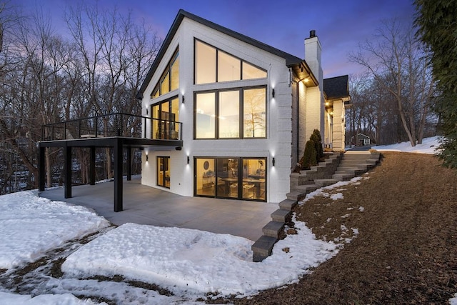 snow covered back of property featuring a patio area, a chimney, stairway, and brick siding